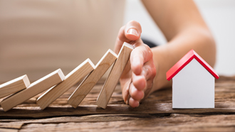 Mão a impedir um dominó de cair sobre uma casa em miniatura a propósito do seguro multirriscos habitação Deco+